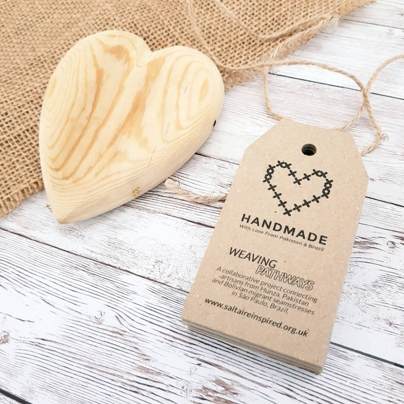 Puede incluir: Un coraz&oacute;n de madera hecho a mano con una etiqueta de papel marr&oacute;n adjunta. La etiqueta tiene un dise&ntilde;o de coraz&oacute;n cosido y el texto "HANDMADE With Love From Pakistan & Brazil WEAVING PATHWAYS A collaborative project connecting artisans from Hunza, Pakistan and Bolivian migrant seamstresses in S&atilde;o Paulo, Brazil, www.saltaireinspired.org.uk".