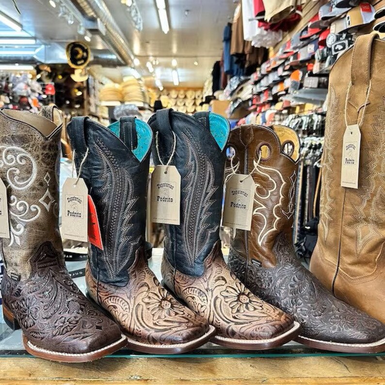 Puede incluir: Cinco pares de botas vaqueras de cuero marr&oacute;n con intrincados bordados florales. Las botas tienen una punta puntiaguda y un tac&oacute;n bajo. Las botas est&aacute;n dispuestas en una fila sobre una superficie de madera. Las botas tienen etiquetas que dicen "Justamente Padrito".