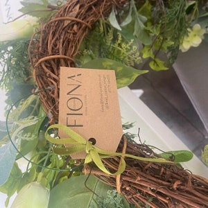 Puede incluir: Una corona verde y marr&oacute;n con una etiqueta marr&oacute;n que dice "Fiona Floral Couture" con informaci&oacute;n de contacto.