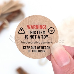 May include: A circular brown sticker with black and red text. The text reads "WARNING! THIS ITEM IS NOT A TOY" and "KEEP OUT OF REACH OF CHILDREN." A warning symbol and the number "14+" are also present.