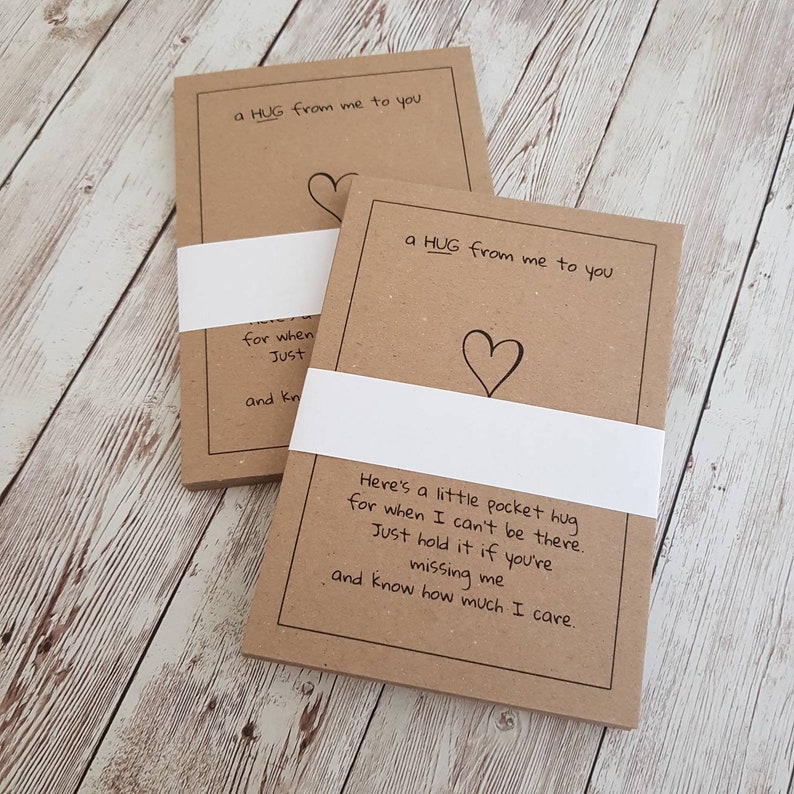 May include: Two brown paper cards with white borders and the text "a HUG from me to you" and a heart drawn on each card. The text on the bottom card reads "Here's a little pocket hug for when I can't be there. Just hold it if you're missing me and know how much I care."