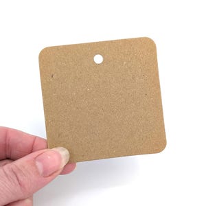 May include: A square, brown kraft paper tag with rounded corners and a small hole at the top. The tag is held by a hand against a white background. The tag is blank, ready for use as a product tag or label.