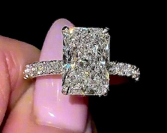 May include: A diamond ring featuring a large, rectangular radiant-cut centre stone. The band is embellished with smaller, sparkling stones. The ring is set against a dark backdrop, accentuating its sparkle. A luxurious piece of jewellery.