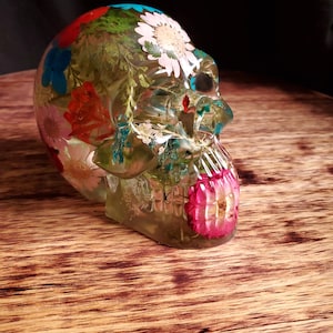 May include: A clear glass skull with dried flowers and greenery embedded inside. The skull is decorated with pink, red, white, and blue flowers.