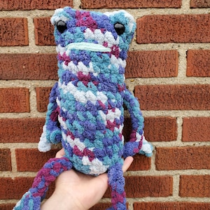 May include: A crocheted frog plush toy with a blue, purple, and white color scheme. The frog has large black eyes and a wide smile.