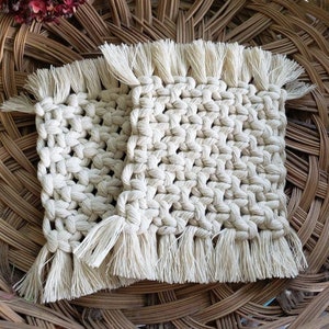 May include: Two square beige crocheted coasters with fringe on all sides. The coasters are sitting on a woven wicker basket.