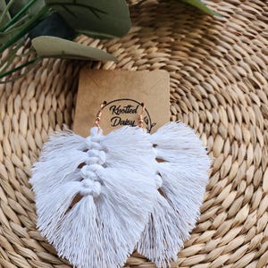 May include: White macrame feather earrings with gold-colored ear wires. The earrings are displayed on a woven, light brown surface with a small brown tag that reads "Knotted Daisy". Green leaves are in the background.
