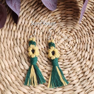 May include: Handmade macrame earrings featuring a sunflower design. The earrings have yellow and green tassels, with a yellow flower at the top. The word "Sunflower" is written in cursive. The earrings are displayed on a woven background.