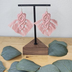 May include: A pair of pink macrame earrings with fringe hanging from a wooden stand. The earrings are displayed on a wooden surface with green leaves.