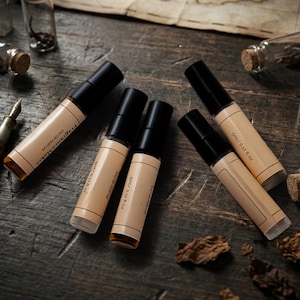 Pode incluir: Uma disposição de frascos de perfume roll-on com tampas pretas e rótulos bege, cada um com a inscrição "Fragrance Alchemy Eau de Parfum" e "5ml / 0.17 fl oz." Pequenos frascos de vidro com rolhas e um mapa vintage.