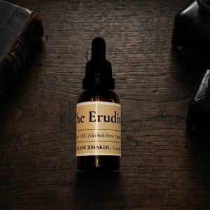 Może przedstawiać: Brązowa szklana butelka z czarną pipetą, z etykietą "The Erudite" na kremowej etykiecie. Butelka stoi na ciemnej drewnianej powierzchni, z tyłu stara książka i kałamarze. Na etykiecie widnieje również napis "Fragrance Oil | Alcohol-Free".