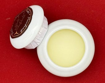 May include: A small white ceramic jar with a lid. The jar contains a pale yellow balm. The lid has a brown label with the text "Handmade" and a stylized tree design.