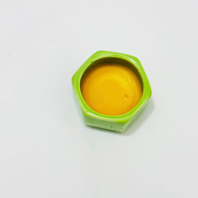 Close-up top view of golden Osmanthus solid perfume balm inside a lime green hexagonal ceramic jar. Rich, creamy beeswax texture with notes of sweet apricot and tea.