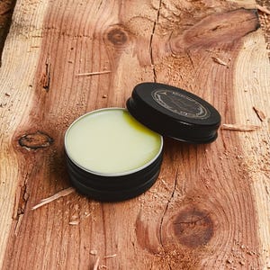 May include: An open, black metal tin filled with a light green balm, next to its lid. The lid is black with a white label. The tin sits on a rough-hewn, reddish-brown wooden surface, with wood shavings scattered around.