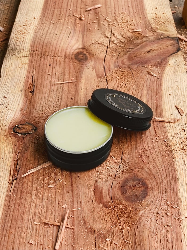 May include: An open, black metal tin filled with a light green balm, next to its lid. The lid is black with a white label. The tin sits on a rough-hewn, reddish-brown wooden surface, with wood shavings scattered around.