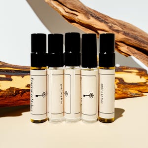 May include: Five glass roll-on perfume bottles with black caps. Each bottle has a cream-coloured label with the text "Fragrance Maker" and a key logo. The bottles are filled with a golden liquid. The background includes a piece of wood and a white surface.
