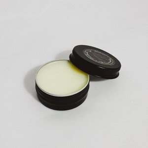 An open tin of Dry Santal solid cologne. A raw woody fragrance with notes of cedar shavings, papyrus, and leather. Industrial and modern unisex scent.