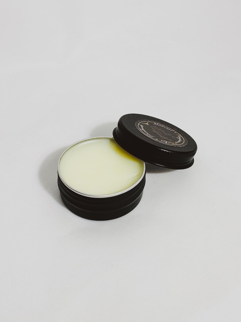An open tin of Dry Santal solid cologne. A raw woody fragrance with notes of cedar shavings, papyrus, and leather. Industrial and modern unisex scent.