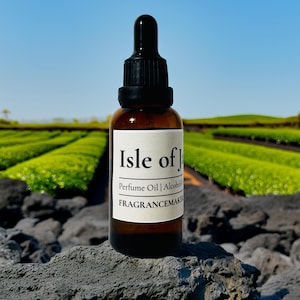 May include: A brown glass perfume oil bottle with a black dropper cap. The label reads "Isle of J" and "Perfume Oil | Alcohol" and "FRAGRANCEMAKER". The bottle sits on a gray rock with a green field and blue sky in the background.