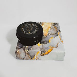 An open tin of Dry Santal solid cologne. A raw woody fragrance with notes of cedar shavings, papyrus, and leather. Industrial and modern unisex scent.