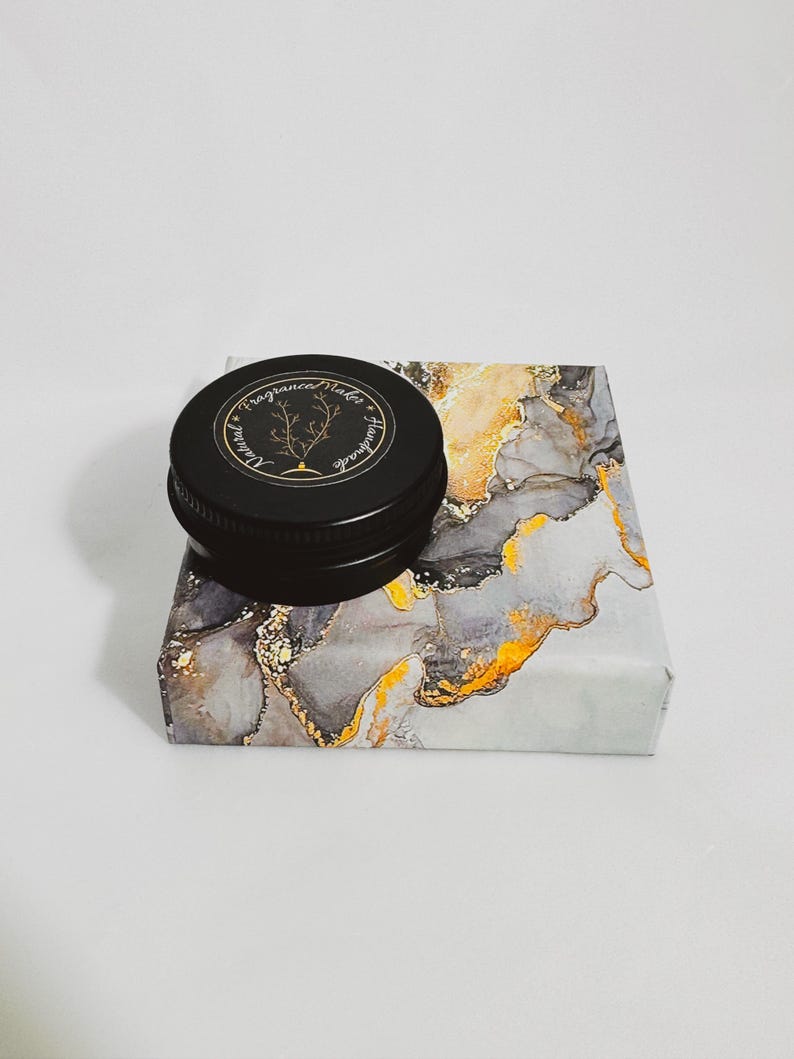 An open tin of Dry Santal solid cologne. A raw woody fragrance with notes of cedar shavings, papyrus, and leather. Industrial and modern unisex scent.