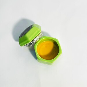 Top-down view of an open Osmanthus solid perfume set. Features a bright green hexagonal ceramic container and a lid with a black botanical label. A sweet, autumnal floral scent.