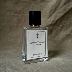 May include: A clear glass perfume bottle with a black cap and a white label. The label reads "Verdant Tincture Eau de Parfum" and "30ml / 1.0 fl oz". The bottle has a gold-colored ring at the base of the cap.