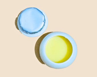 May include: A small, round, light blue container with a yellow balm inside. The container has a separate light blue lid.