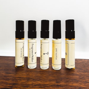 Może przedstawiać: Pięć małych butelek perfum z czarnymi zakrętkami. Etykiety zawierają napisy "FragranceMaker" i "Eau de Parfum". Jedna butelka jest oznaczona jako "5ml / 0.17 fl oz". Butelki ustawione są na ciemnobrązowej drewnianej powierzchni.