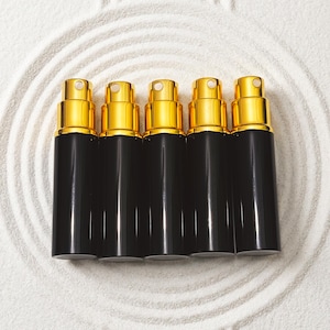 Pode incluir: Cinco frascos de spray pretos com tampas douradas estão alinhados. As garrafas cilíndricas e elegantes se destacam em um fundo branco texturizado com padrões circulares concêntricos. As tampas douradas têm um acabamento brilhante.