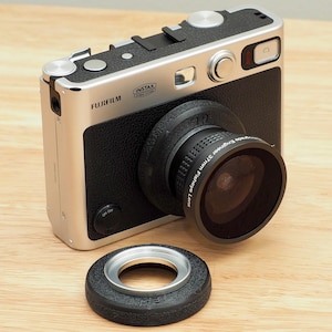May include: Silver and black Fujifilm Instax camera with a 37mm fisheye lens attached. The camera has a black leather-like grip and silver accents. An additional black lens ring sits in front of the camera on a wooden surface.