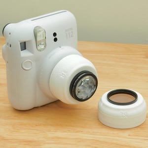 Può includere: Una fotocamera istantanea Instax i12 bianca con un filtro per lenti bianco attaccato e un filtro separato. La fotocamera e i filtri sono su una superficie di legno. Il filtro dell'obiettivo ha un anello nero e una lente trasparente sfaccettata.