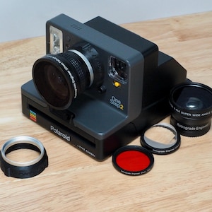 Puede incluir: Una cámara Polaroid OneStep 2 negra con un objetivo de ojo de pez. La cámara está rodeada de varios accesorios de objetivo, incluyendo un objetivo gran angular, un filtro rojo y un filtro transparente.