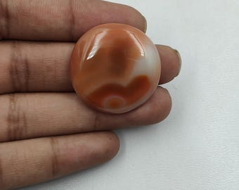 May include: A close-up of a smooth, oval-shaped agate cabochon with a swirling pattern of orange, white, and brown.