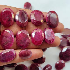 May include: A selection of oval-cut, deep red rubies. The gemstones are held in a hand, with several loose rubies scattered around. The rubies have a faceted surface, catching the light and displaying their rich colour.