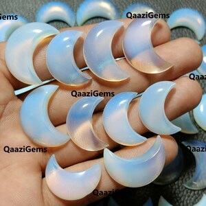 Puede incluir: Una colección de cabujones de ópalo blanco iridiscente en forma de media luna. Los cabujones son lisos y pulidos, con una variedad de tamaños y formas. Los cabujones están dispuestos en una mano, con las puntas de las medias lunas mirando al espectador.