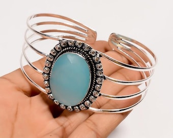 May include: Silver cuff bracelet with a large, oval, pale blue gemstone. The gemstone is set in a detailed, floral-patterned bezel. The bracelet has multiple parallel bands and a hinged design.