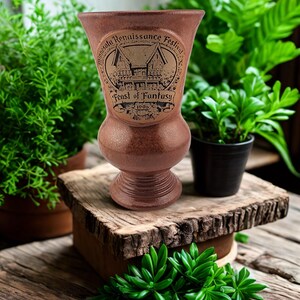May include: A brown ceramic goblet with a flared rim and a pedestal base. The front features a circular embossed design with the text Minnesota Renaissance Festival, Feast of Fantasy, and the year 1996, depicting a timber-framed building.