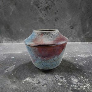 May include: A ceramic vase with a wide, angled shoulder and a tapered base. The surface features a textured, horizontal ribbed pattern with a mottled glaze in shades of deep red, muted blue, and charcoal gray, creating a weathered appearance.