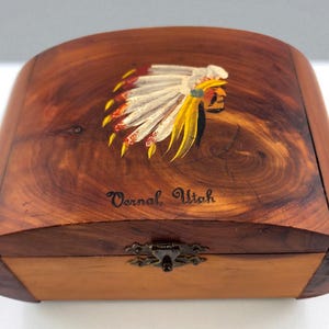 May include: A wooden box with a curved top and a hinged front. The top is decorated with a painted Native American headdress in white, yellow, red, and black. The text "Vernal, Utah" is written on the box.