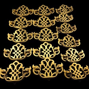 May include: A collection of ornate, gold-colored drawer pulls arranged against a black background. Each pull features a decorative, openwork design with curved handles and small, round accents. The pulls are likely for furniture restoration or decorative purposes.