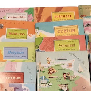 May include: A collection of vintage travel books from the American Geographical Society's "Around the World Program." The covers feature colorful illustrations and country names like Mexico, Portugal, and Ceylon. The books are stacked together.