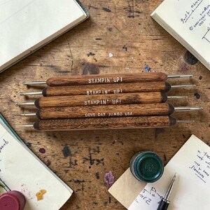 May include: A collection of wooden Stampin' Up! tools with metal tips, labeled "DOVE D47 JUMBO USA." The tools are arranged on a wooden surface, alongside stationery and ink. The tools are used for crafting and scrapbooking.