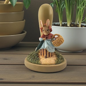 May include: A decorative figurine of a rabbit dressed in a red cape and blue dress, holding a basket. The rabbit stands on a small, oval base with a path design. The figurine is set against a wooden surface.