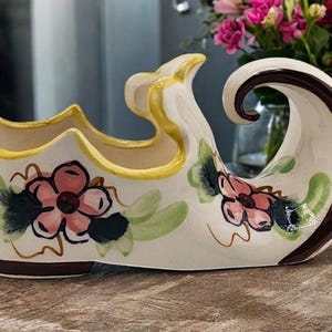 May include: A white ceramic shoe-shaped planter with a brown rim and floral designs. The planter has a yellow rim and pink flowers with green leaves.