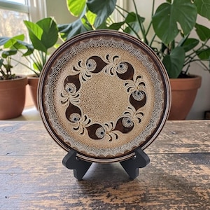May include: A round ceramic plate featuring a speckled tan center and a dark brown border. The plate is decorated with a circular pattern of eight dark brown teardrop-shaped motifs, each containing a small black center and white line details.