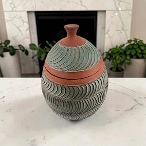 May include: A round ceramic jar with a lid, featuring a terracotta-colored base and a textured, sage-green surface with carved, repeating wave patterns. The lid has a small, knob-like handle at the top, and the overall design is symmetrical.