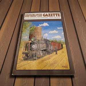 May include: A vintage "Narrow Gauge and Short Line Gazette" magazine. The cover features a detailed illustration of a steam train, water tower, and boxcars in a rural setting. The magazine is set on a wooden surface.