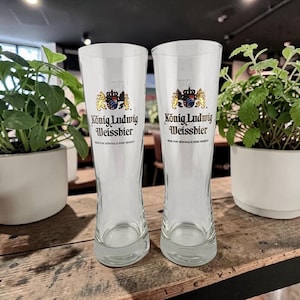 May include: Two clear, tall beer glasses with the "König Ludwig Weissbier" logo. The logo features a gold crown and crest. The glasses are on a wooden surface, with potted plants in the background.