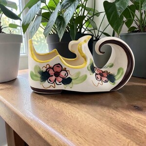May include: A vintage ceramic shoe-shaped planter. The planter is off-white with yellow trim and brown accents. It features pink floral designs with green leaves. The planter is sitting on a wooden surface.